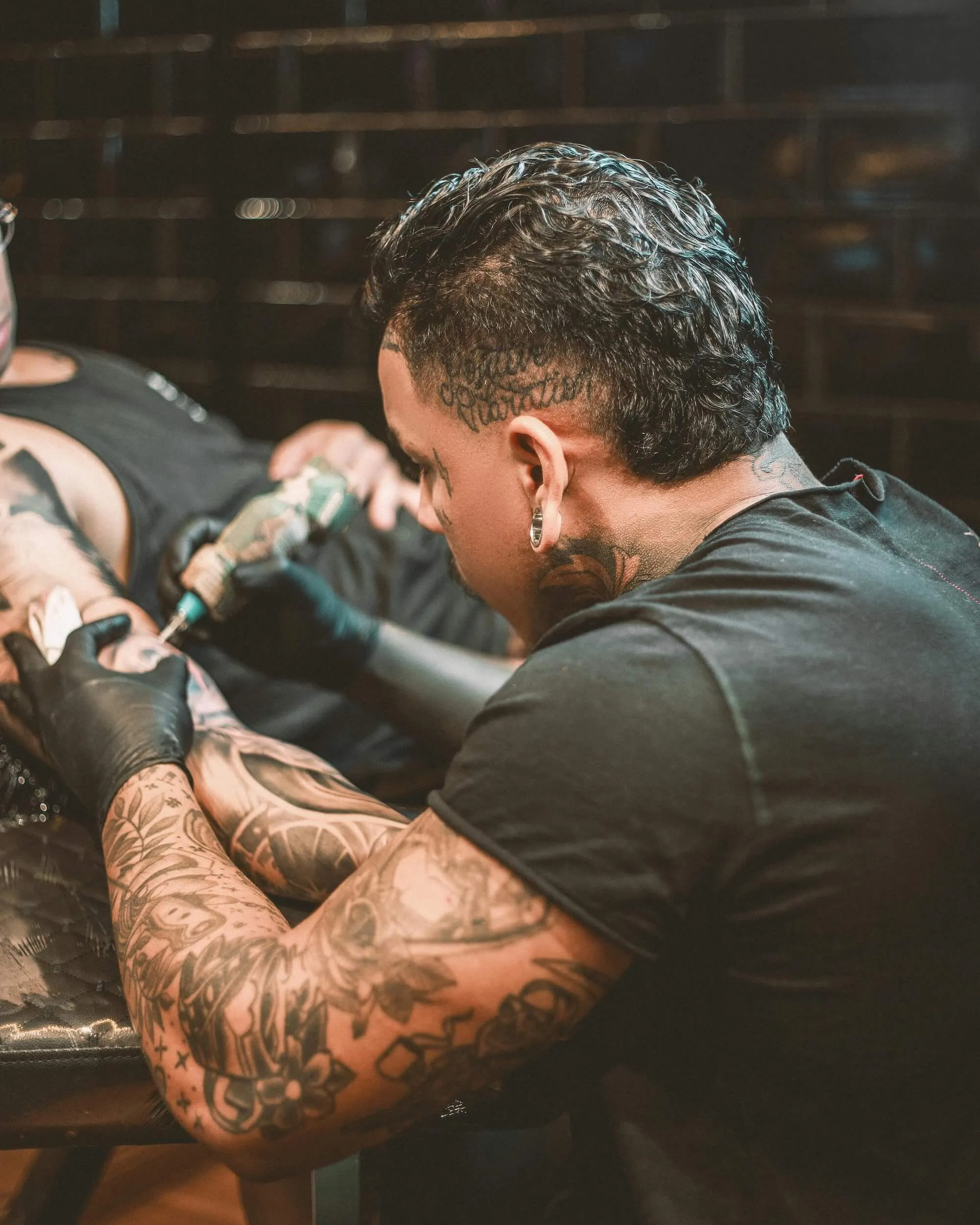 Tattoo artist with sleeve tattoos and ear piercing working on a client's arm while wearing black gloves.