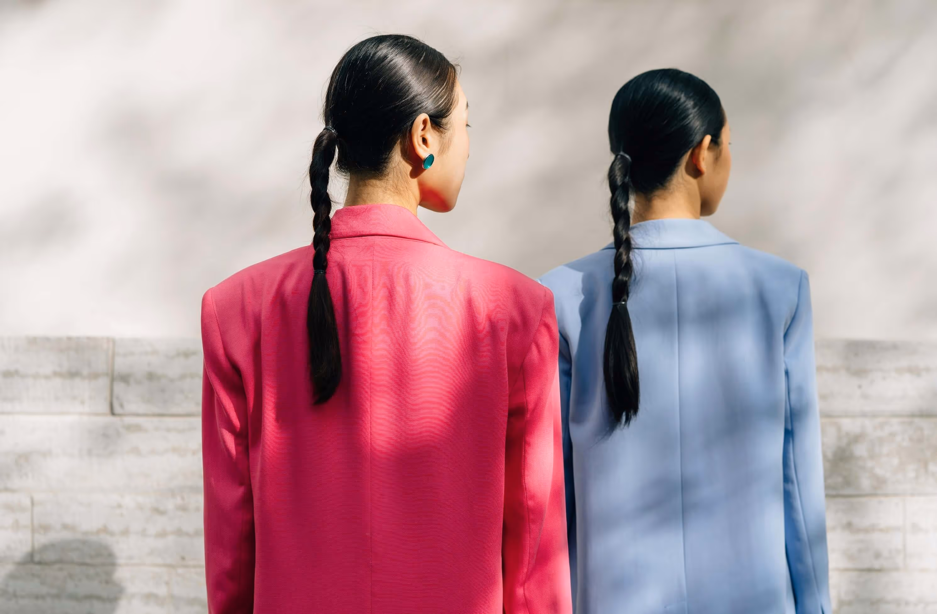 Two women with braided hair standing back to back wearing a pink blazer and a light blue blazer.