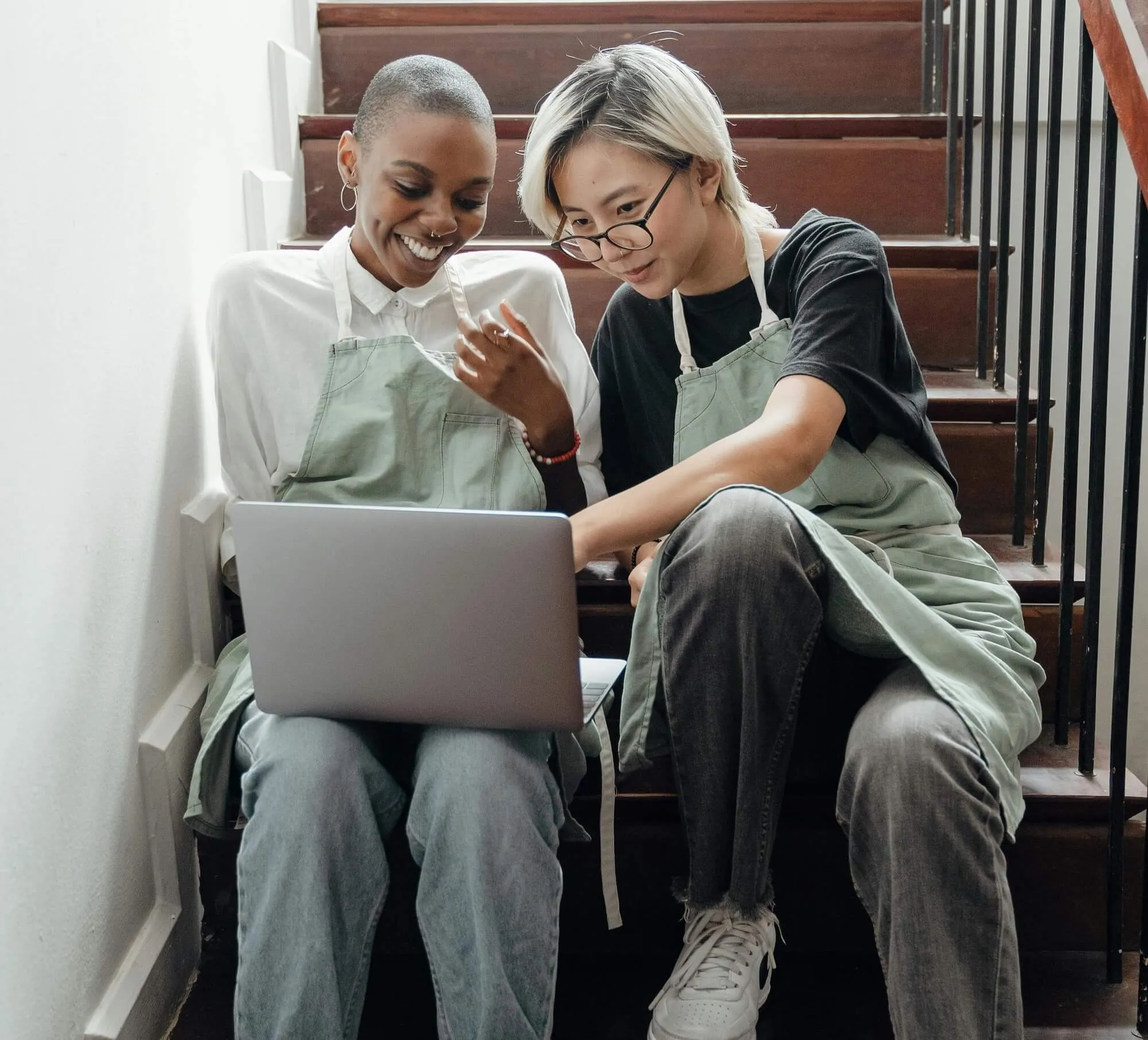 Two people wearing aprons sitting on stairs, looking at a laptop and smiling.