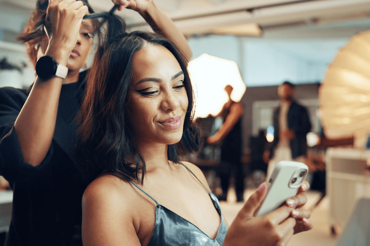 A hair salon client browsing social media on her phone while a hairstylist works on her hair in a modern luxury salon