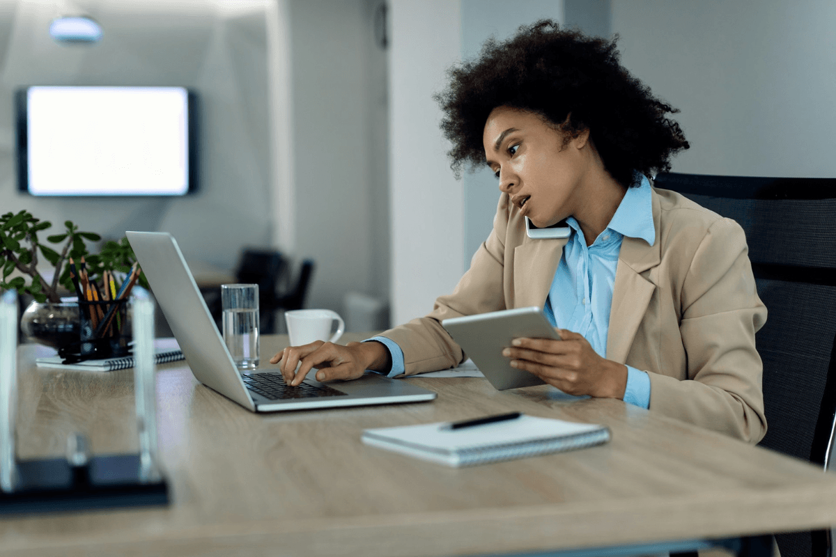 A busy woman multitasking with a phone, laptop, and tablet at her desk, illustrating how easily appointments can be forgotten in a hectic schedule.