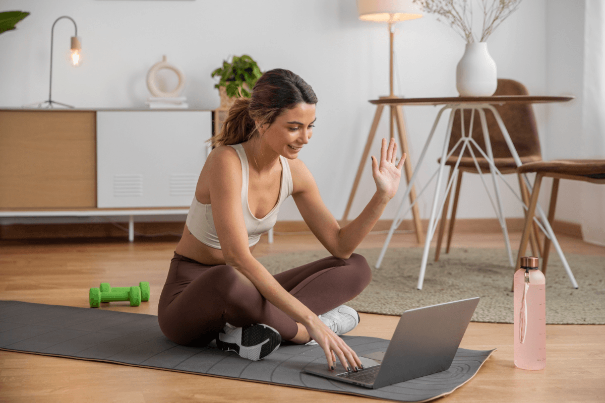 Pilates instructor teaching an online mat class from home, waving at laptop camera with dumbbells on the mat