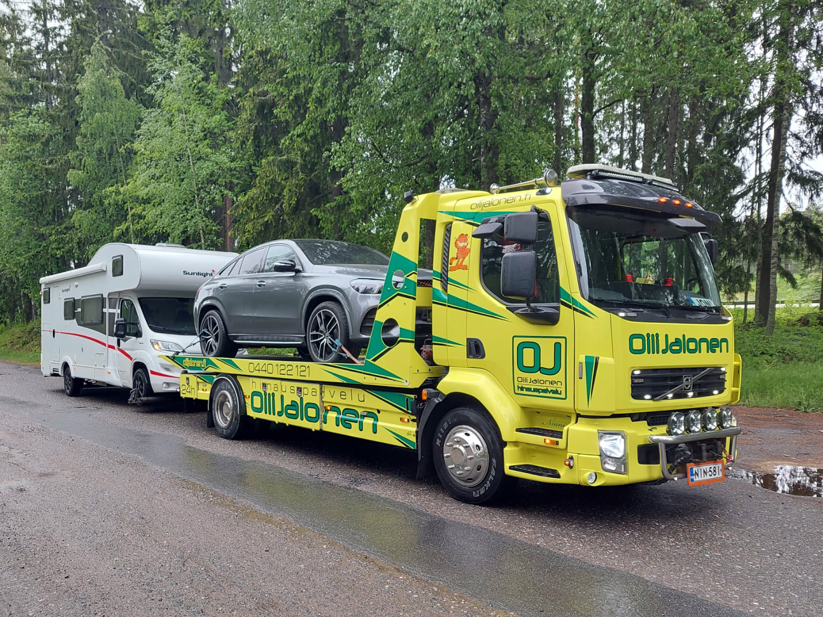 Oili Jalosen keltainen hinausauto kuljettaa harmaata autoa lavalla ja hinaa matkailuautoa.