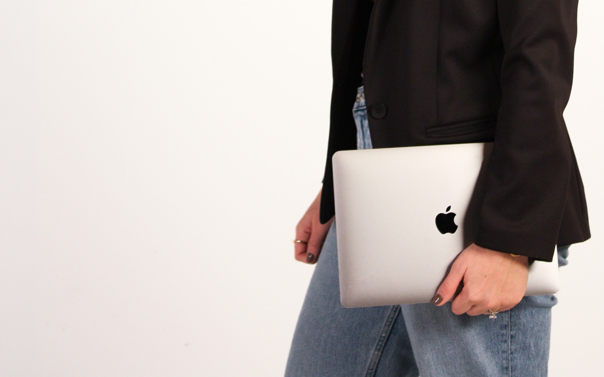 Person wearing a black blazer and blue jeans holding a closed silver Apple MacBook laptop against their hip with a plain white background.