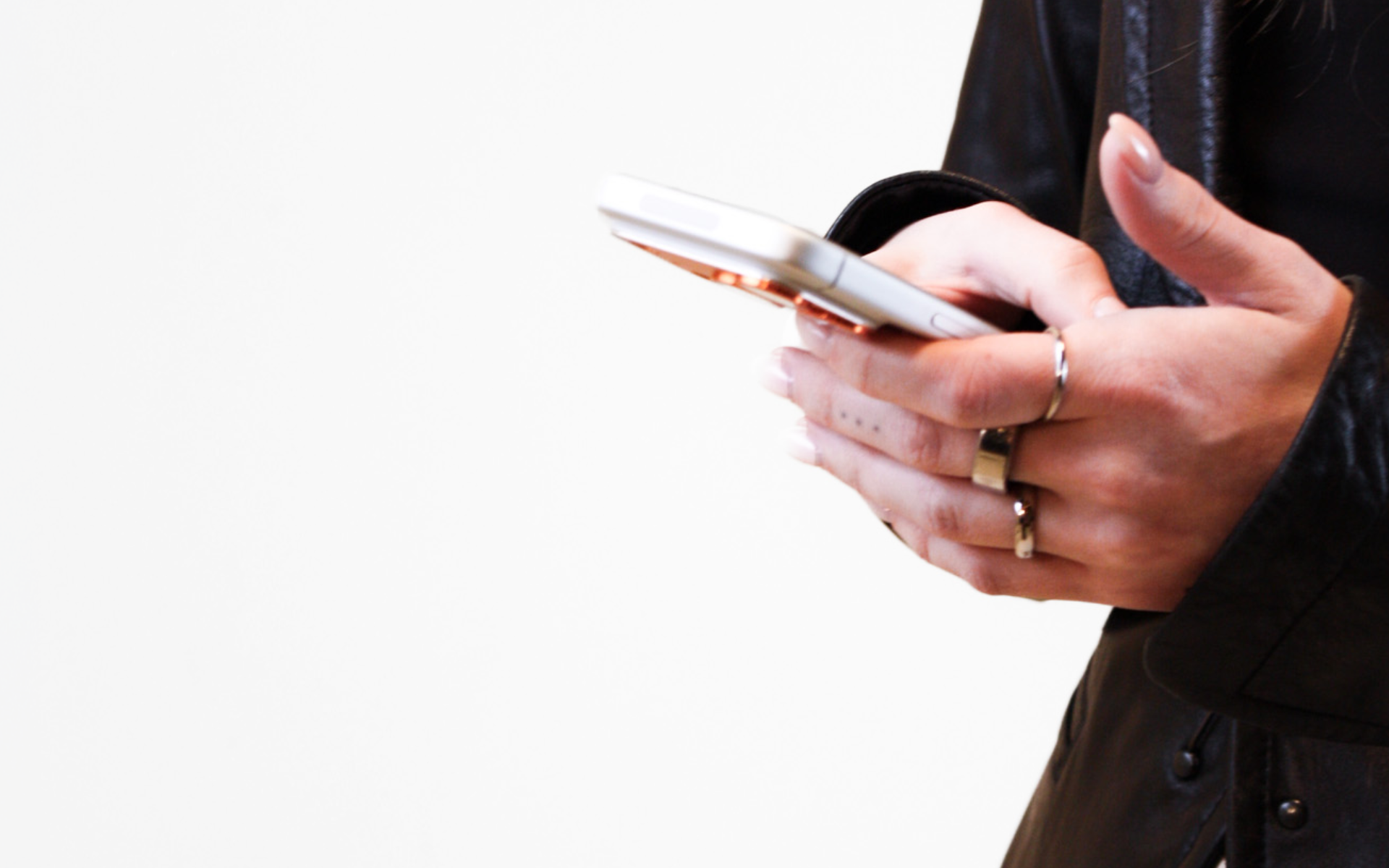 Person wearing a black leather jacket holding and using a smartphone with multiple rings on their fingers.