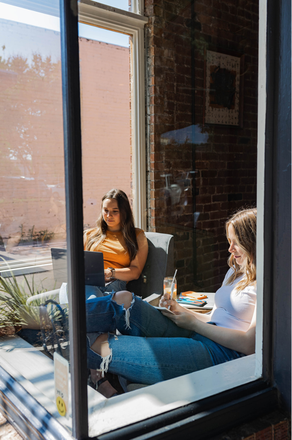 Two women sit by large window in casual clothes working on a laptop and writing notes with a drink nearby.