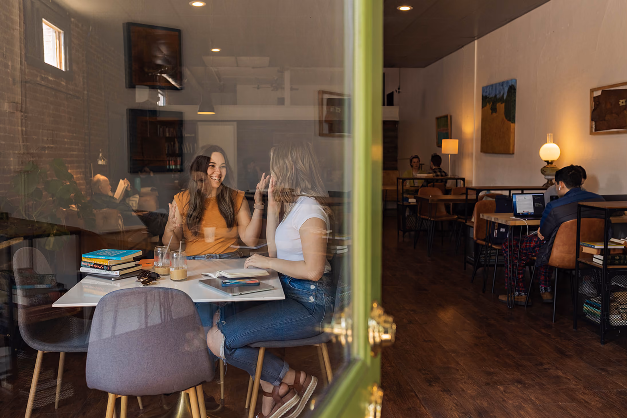 Two women laughing and talking at a table with books and drinks inside a cozy cafe.