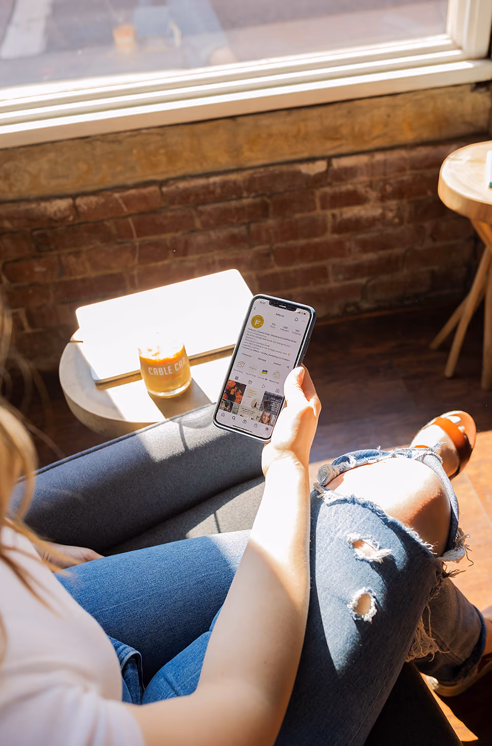 Person wearing ripped jeans holds a smartphone showing an Instagram profile, sitting by a brick wall.