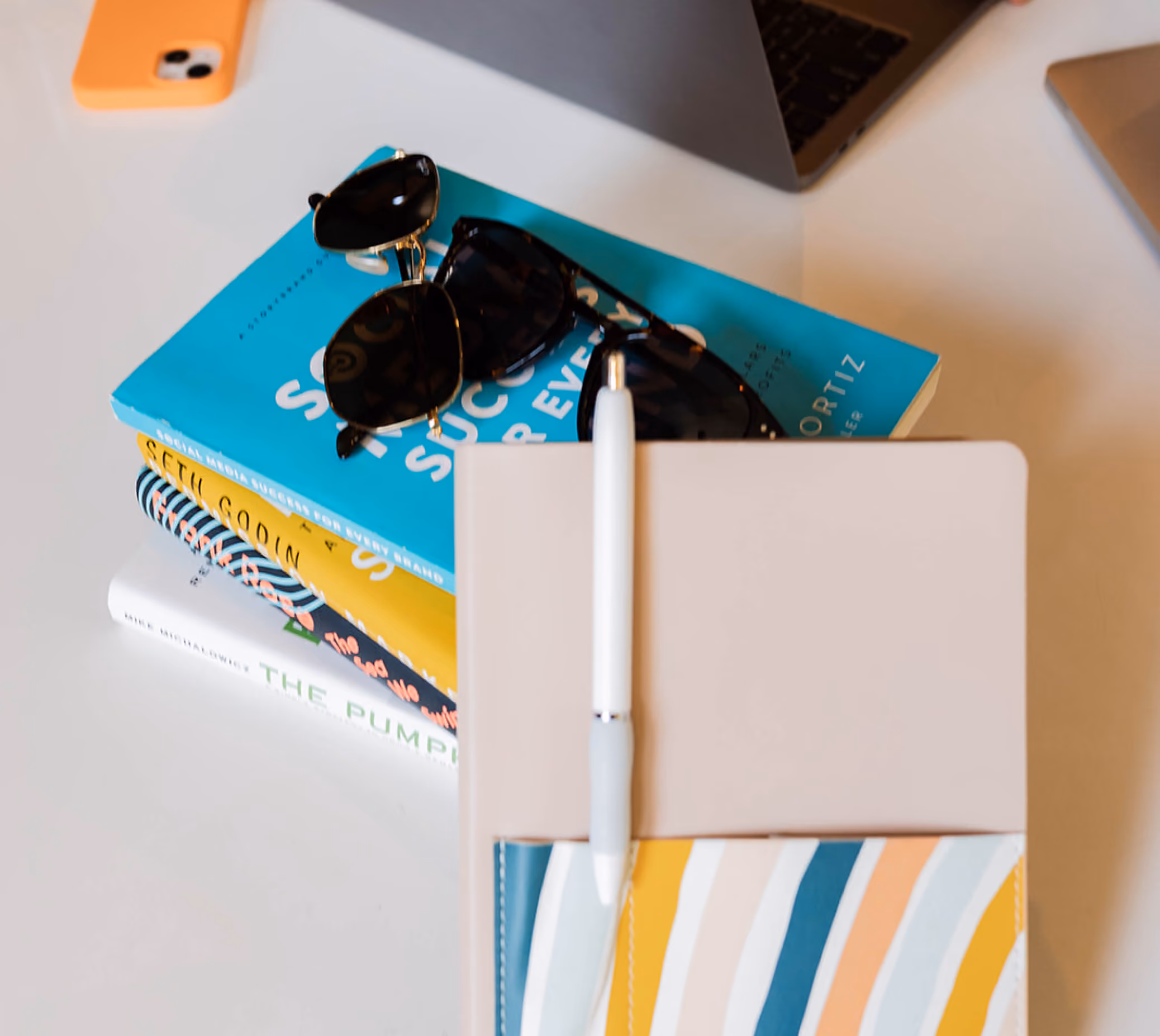 Stack of colorful books topped with two pairs of sunglasses and white pen on a notebook on white table.