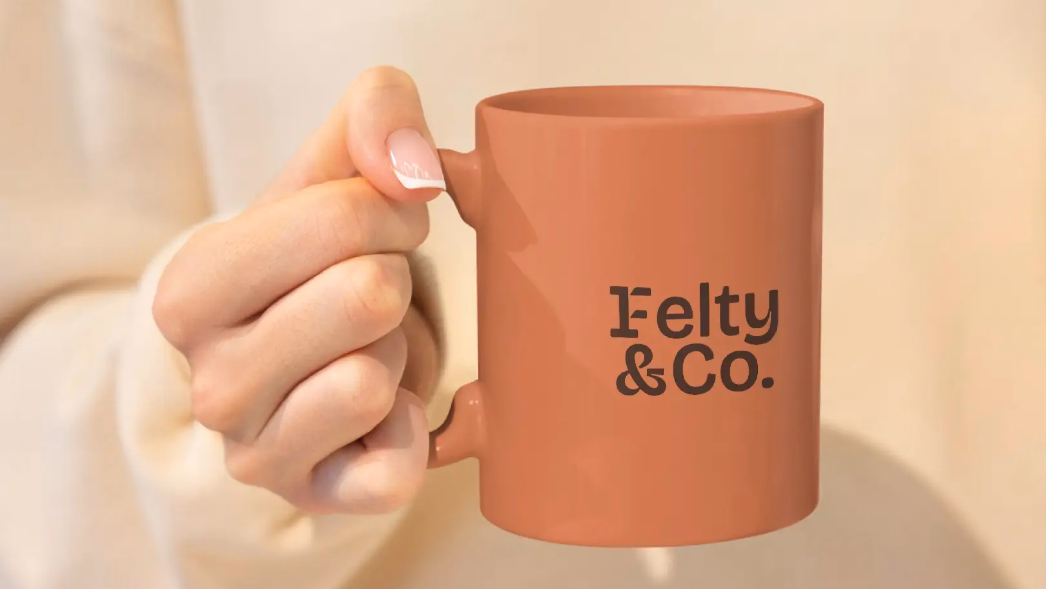 Hand holding a terracotta mug with Felty & Co. printed on it against a beige background.