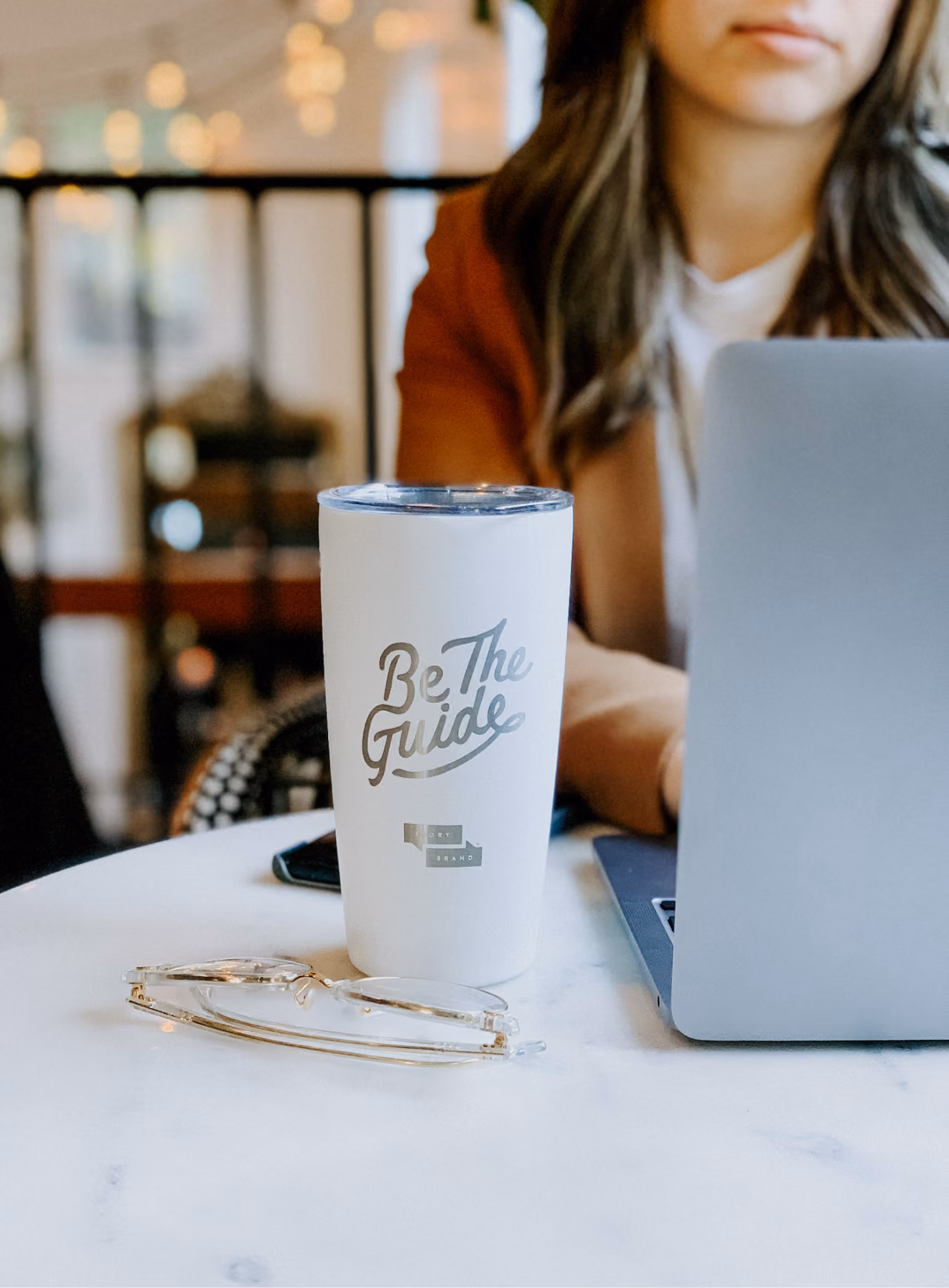 White tumbler with "Be The Guide" text sits on table near eyeglasses and laptop with person in background.