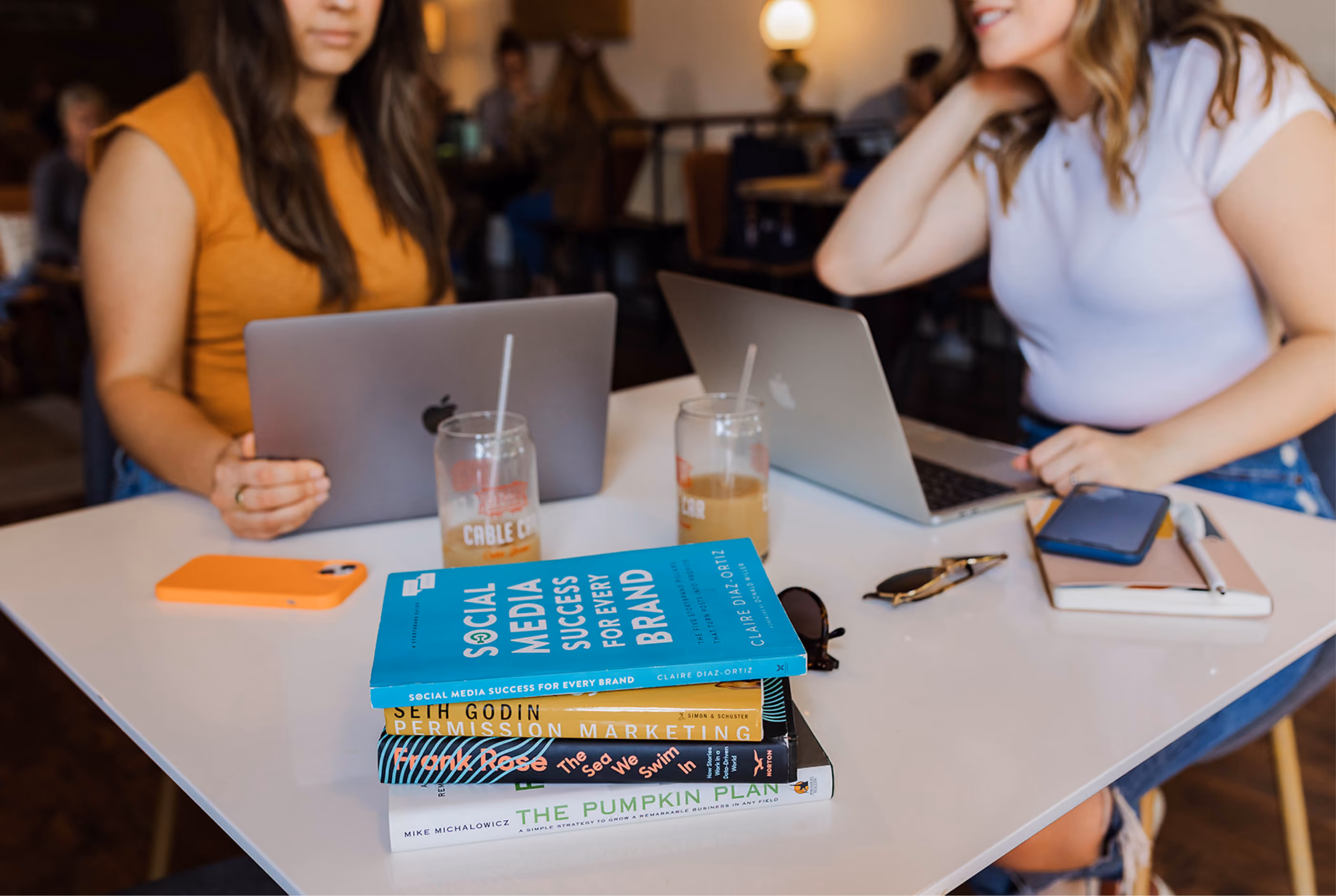 Two women using laptops at a table with a stack of marketing books, drinks, and phones.