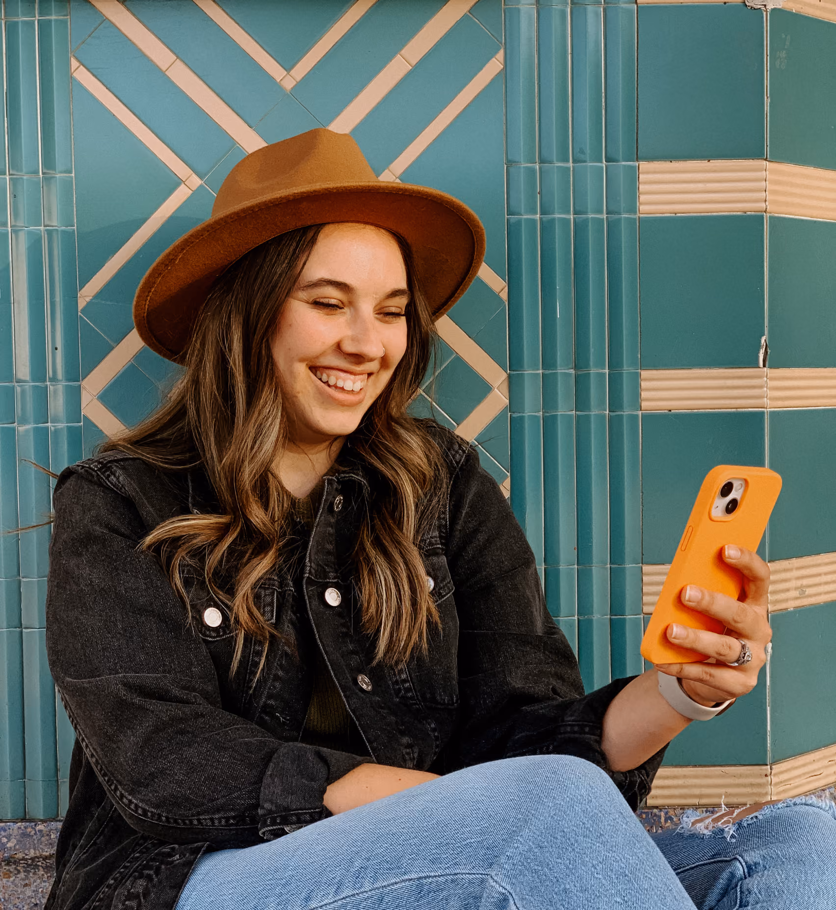 Smiling woman in brown hat and black jacket looking at phone with orange case against teal tiled wall.