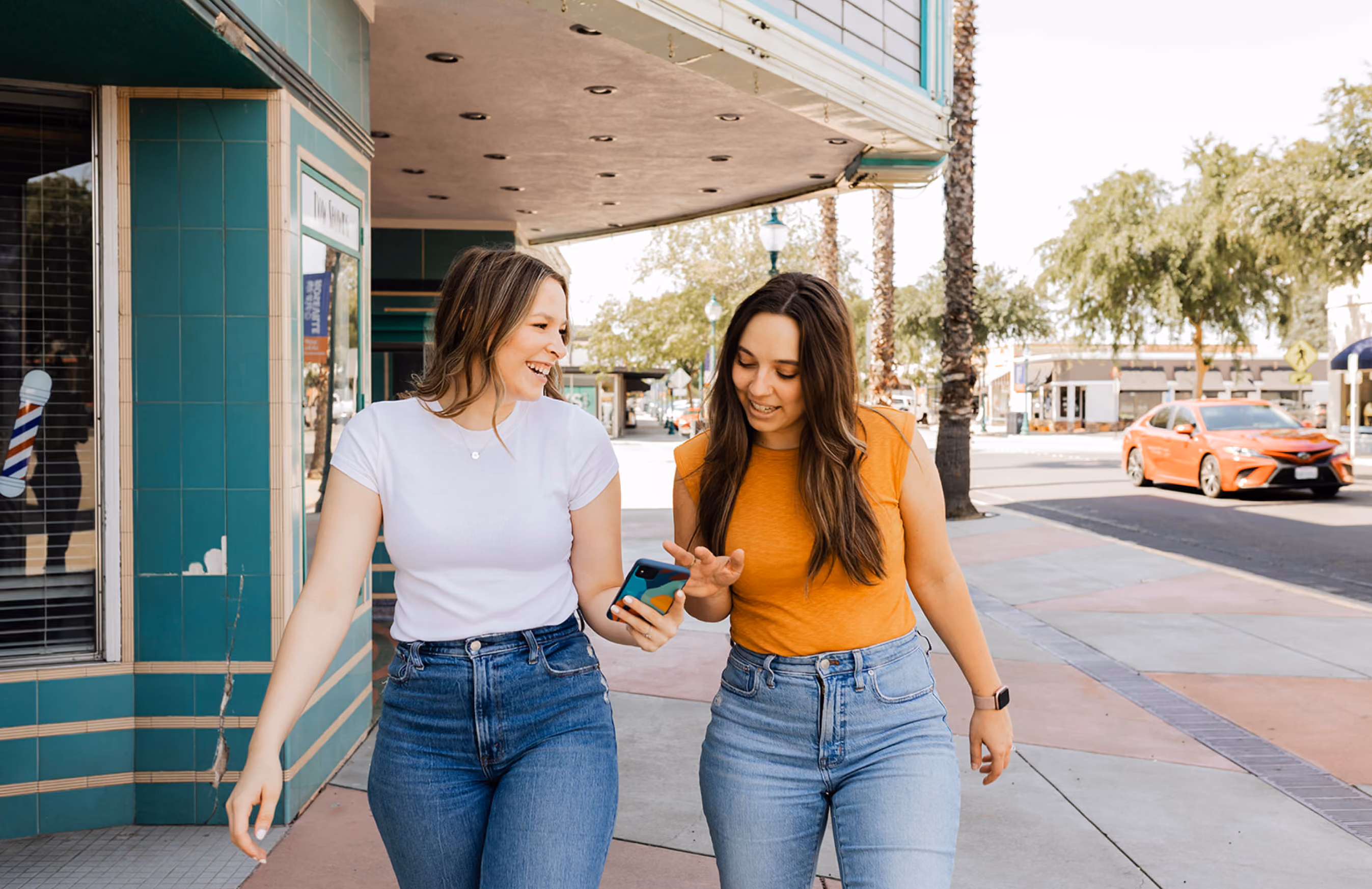 Two women walking on a sidewalk, smiling and looking at a smartphone together.