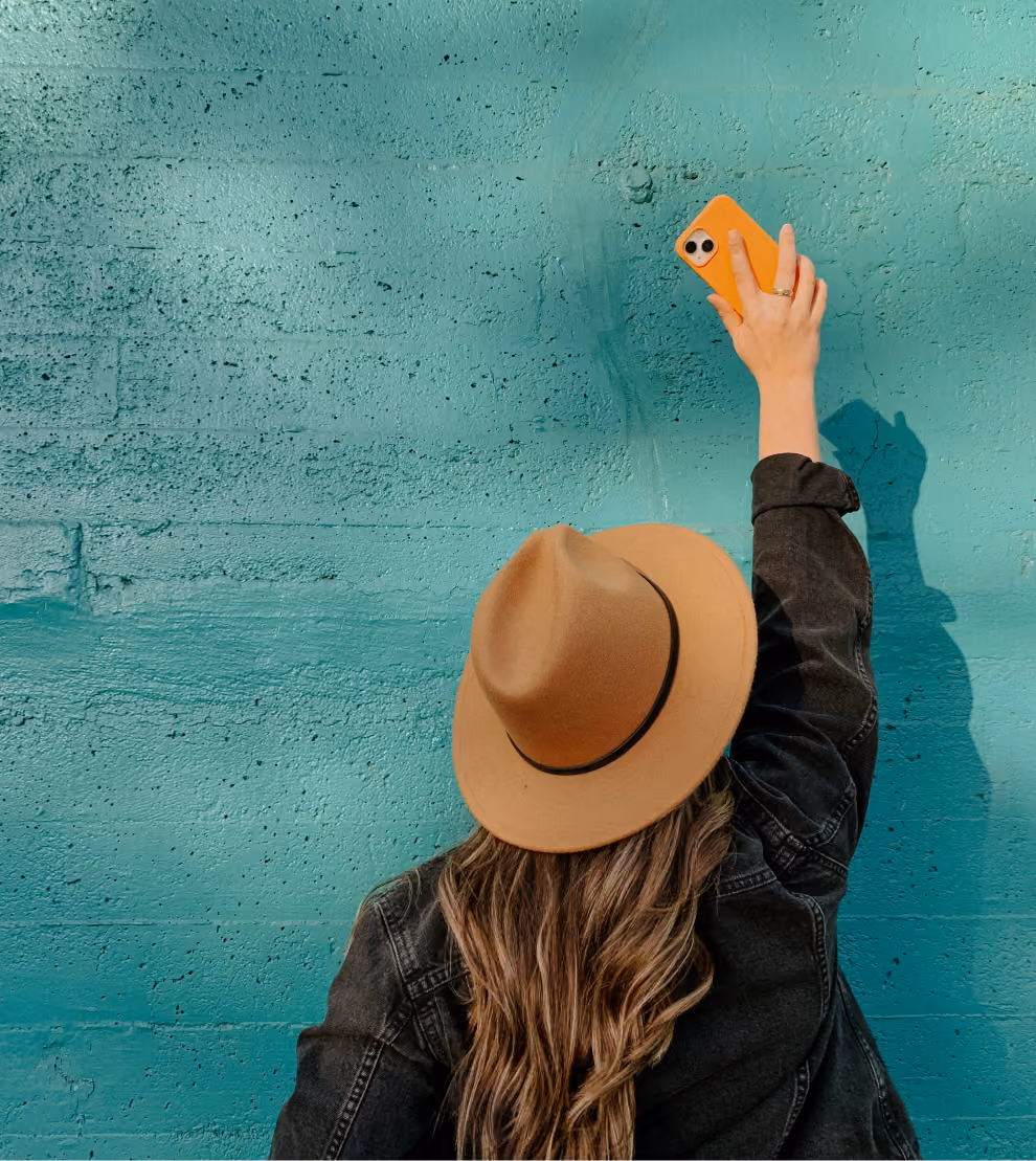 Person in tan hat and dark jacket holds orange phone up against a turquoise textured wall.