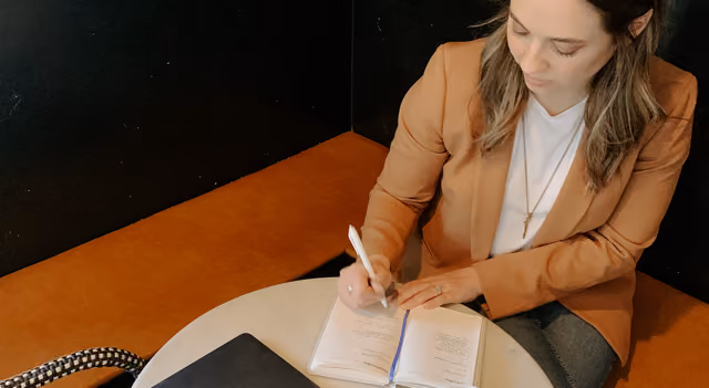 Woman in brown blazer writing in a notebook at a small round table with a laptop nearby.