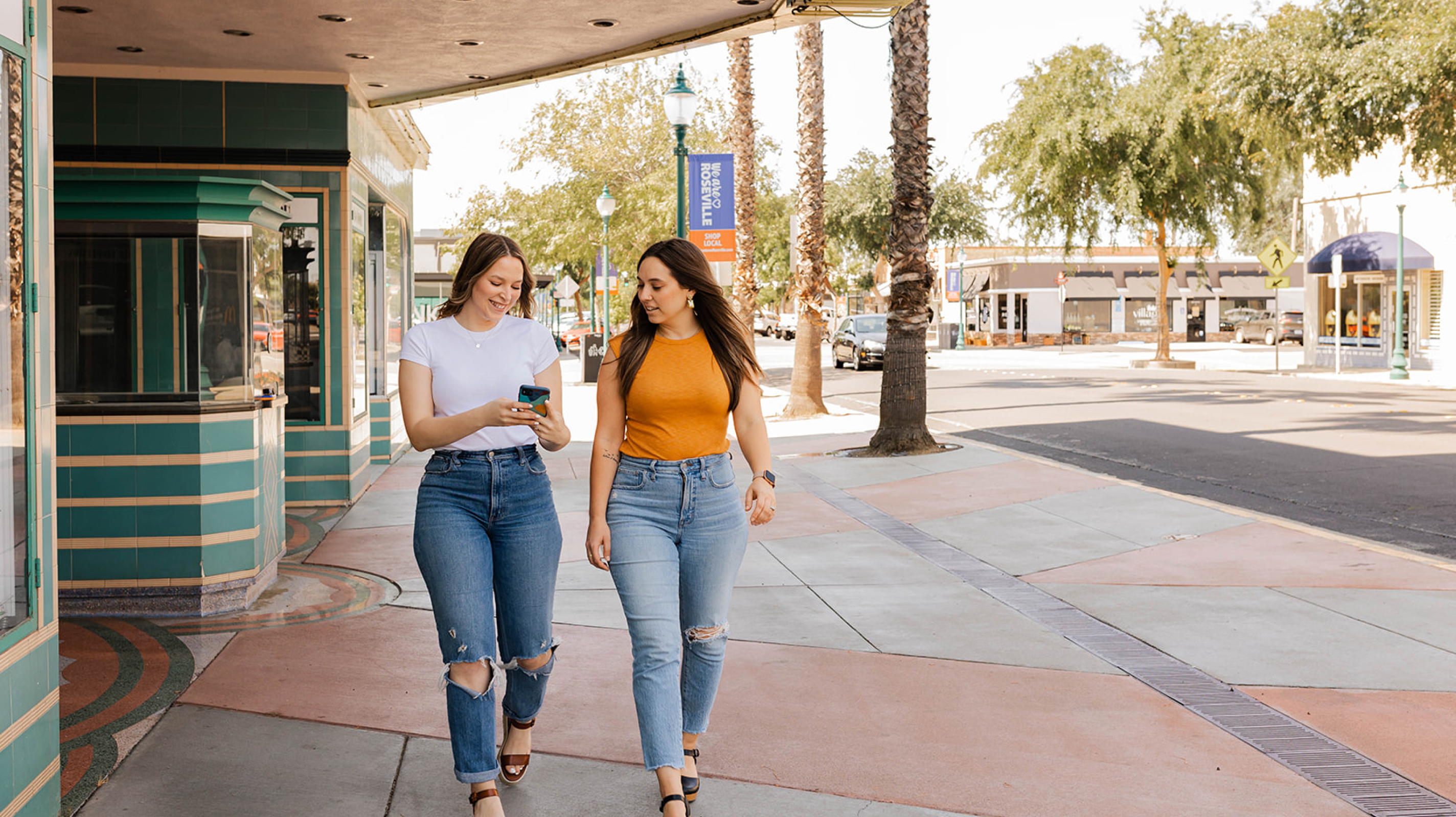 Two women walking on a sunny street, one looking at her phone, the other beside her.