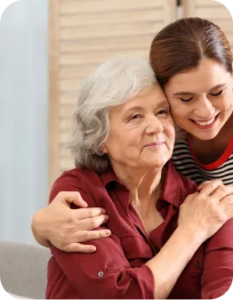 Une jeune femme souriante enlace affectueusement une femme âgée assise à côté d’elle.