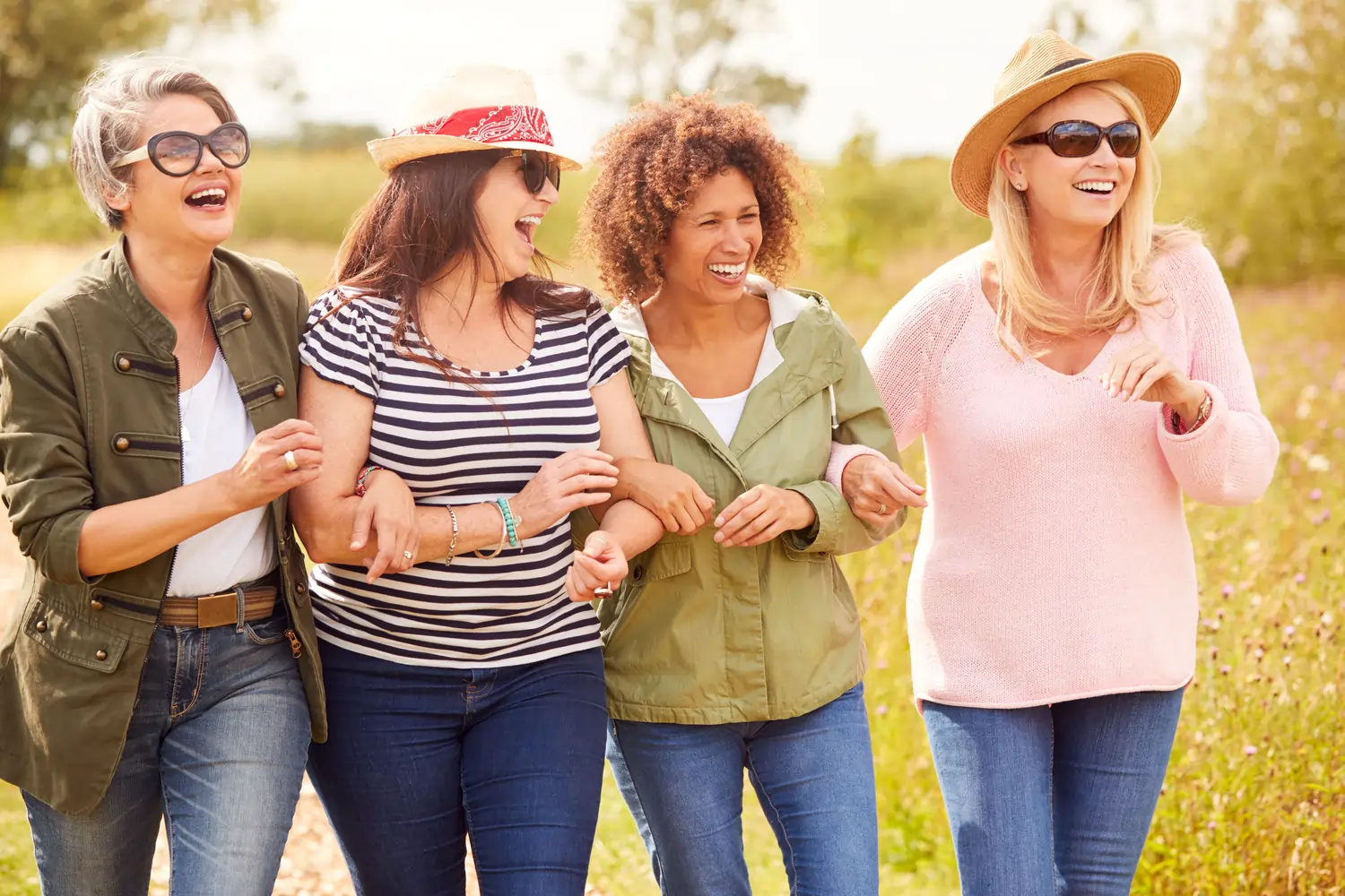 Quatre femmes souriantes marchant bras dessus bras dessous dans un champ ensoleillé.
