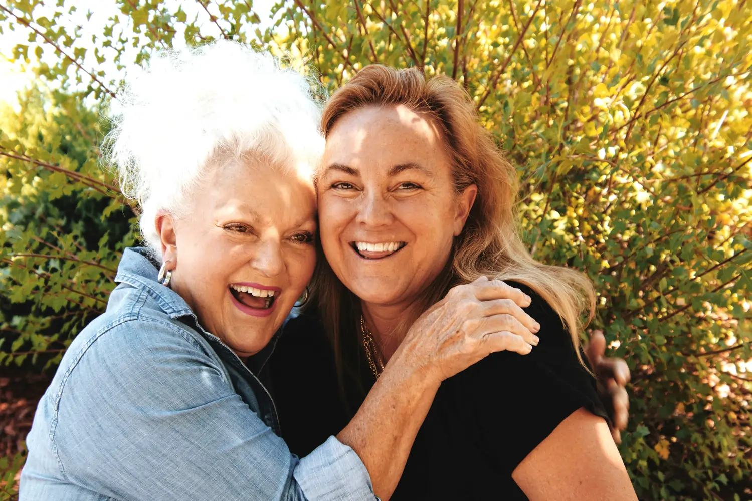 Deux femmes âgées souriantes s'enlaçant joyeusement devant un buisson feuillu au soleil.
