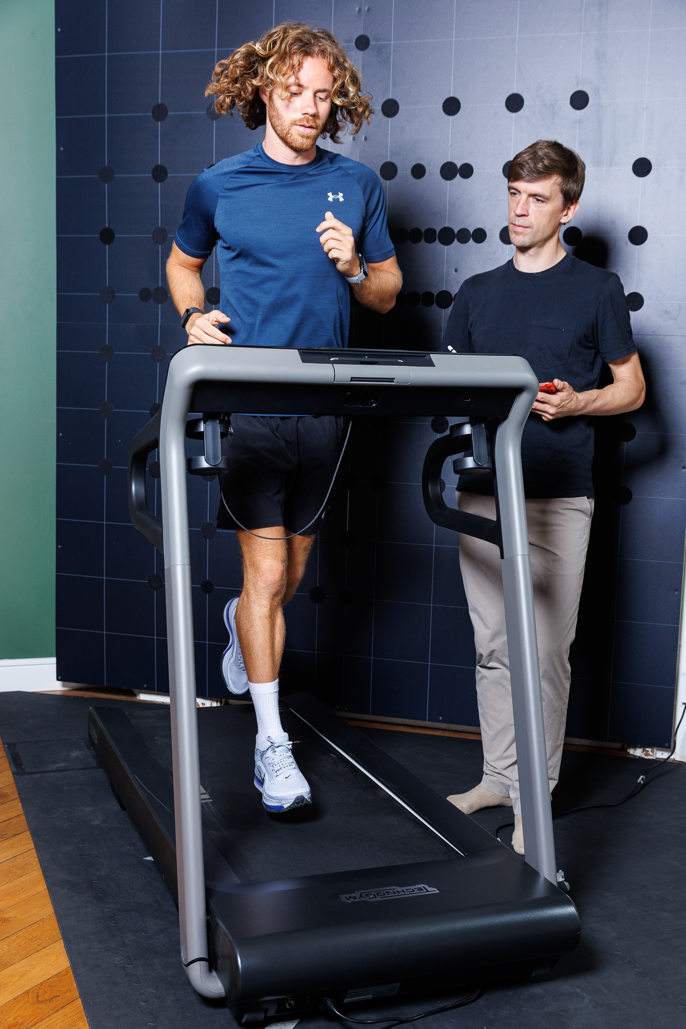 Un homme court sur un tapis de course sous la supervision d’un professionnel de santé ou d’un coach, qui se tient à côté de lui pour observer et mesurer la performance, dans un environnement de cabinet ou de centre de rééducation.