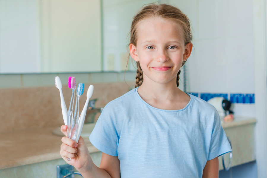 Smiling girl with braided hair holds four toothbrushes in a clean bathroom, highlighting the importance of choosing the right toothbrush for kids.