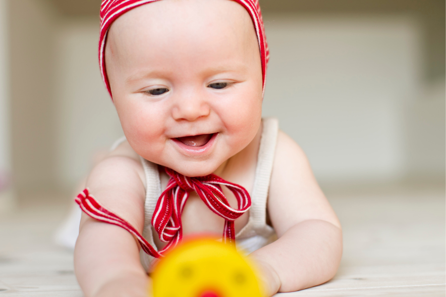 Smiling baby in a red-striped bonnet reaches for a yellow toy while lying on the floor, representing early childhood development and infant dental care awareness