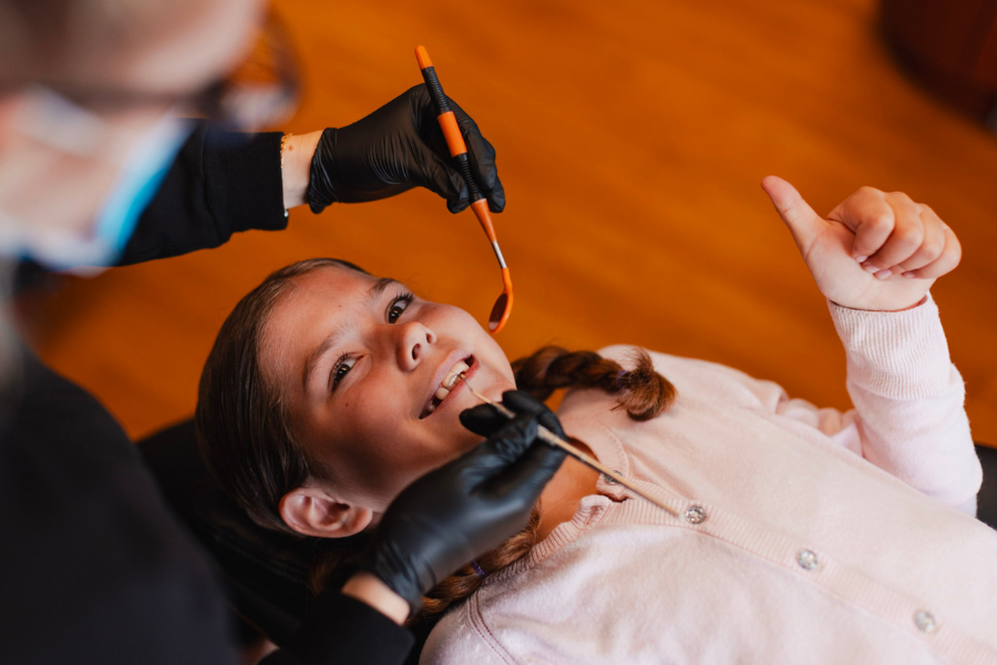 Young girl smiling during a pediatric dental exam while a dentist uses dental tools for a routine teeth cleaning and checkup.