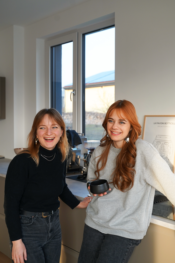 Two young women laughing and chatting in a kitchen, one holding a black cup and both dressed casually.