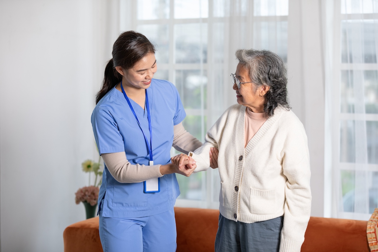 Pflegerin in blauer Uniform unterstützt ältere Frau mit Gehhilfe in hellem Wohnraum.