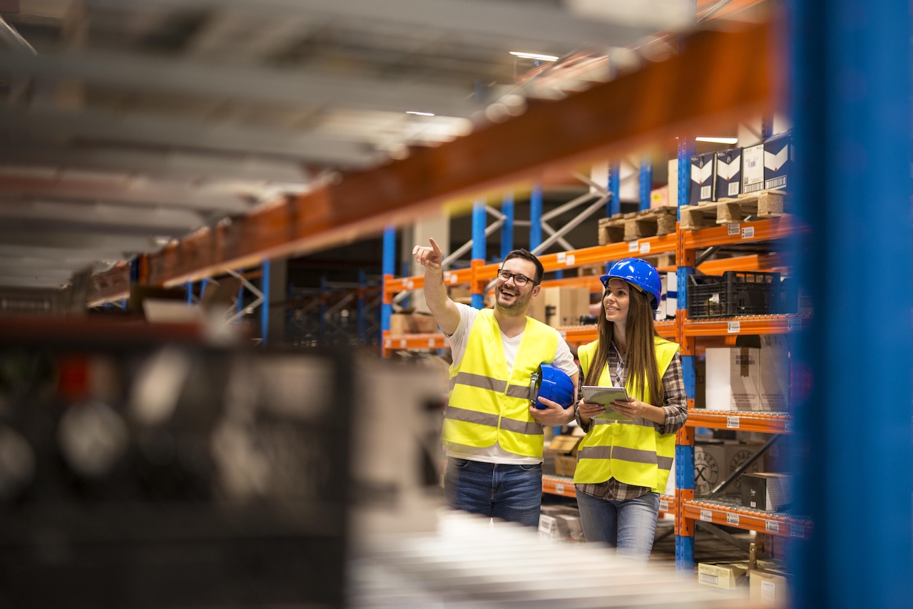 Zwei Lagerarbeiter in Schutzkleidung – Mann und Frau mit Warnwesten und Schutzhelmen – inspizieren ein Lagerhaus.