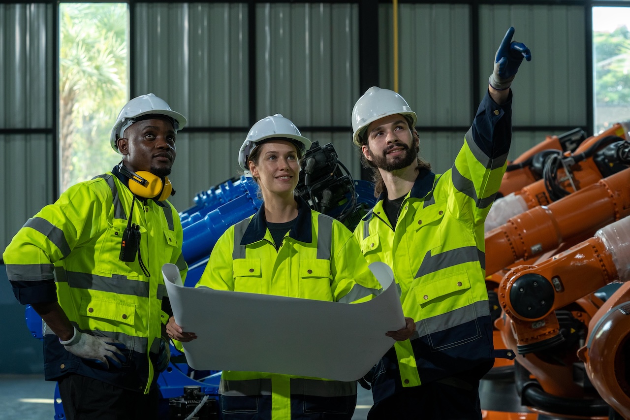 Drei Industriearbeiter in leuchtend gelben Warnwesten, weißen Schutzhelmen und Arbeitshandschuhen inspizieren gemeinsam Baupläne in einer Fabrikhalle. 