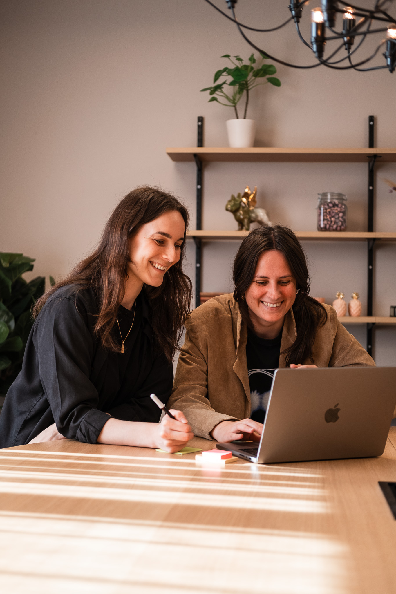 Två kvinnor sitter vid ett bord och ler medan de tittar på en bärbar dator tillsammans, omgivna av hyllor med krukväxter och dekorationer.