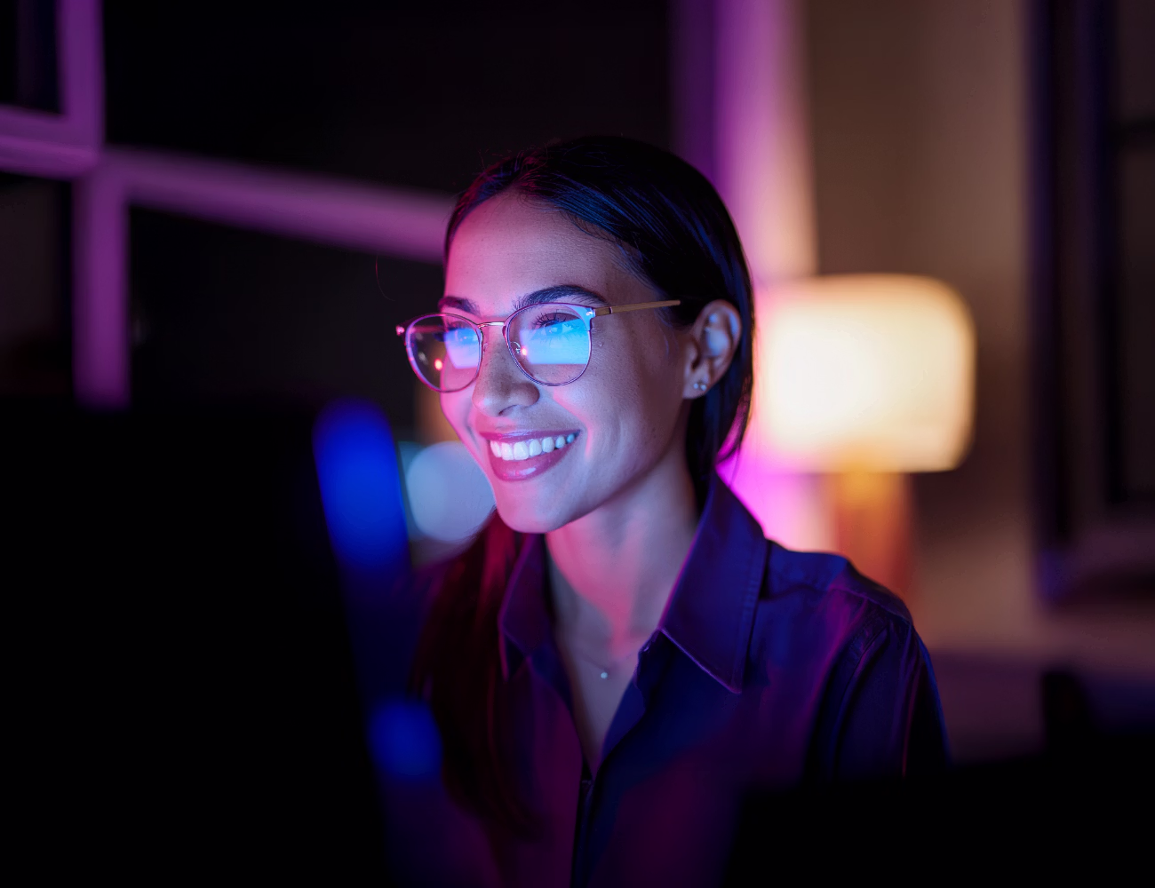 Woman wearing glasses smiling while using a computer, illuminated by blue and pink light, representing AI-assisted research and digital technology.