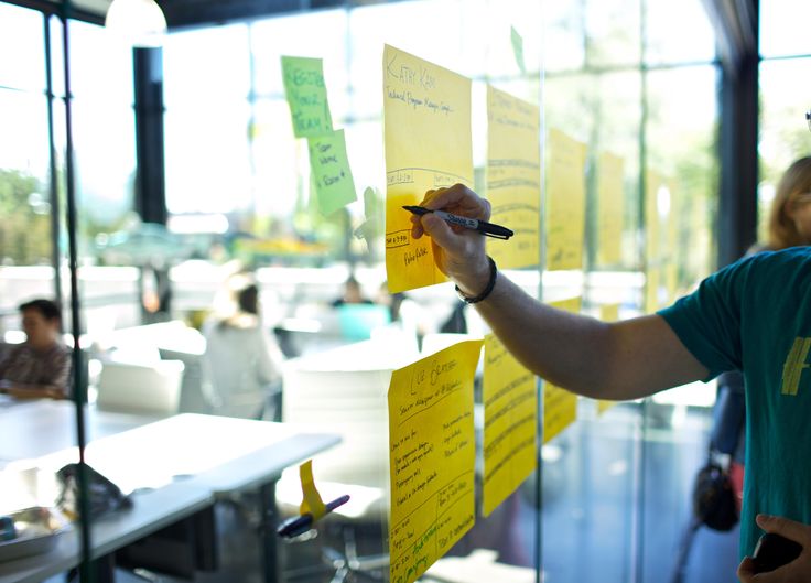 Ricercatore che scrive su un post-it attaccato ad una parete di vetro. Persone che lavorano, fuori fuoco sullo sfondo 