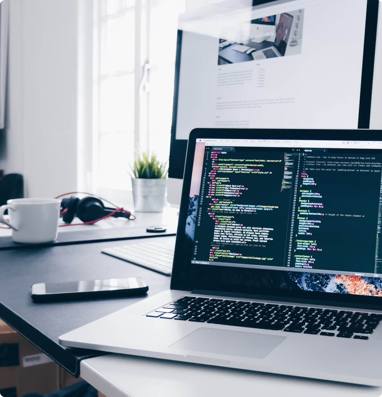 Developer workspace showing laptop with code editor open, coffee cup, smartphone, and development documentation on a clean white desk