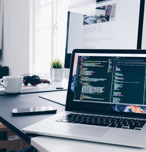 Developer workspace showing laptop with code editor open, coffee cup, smartphone, and development documentation on a clean white desk
