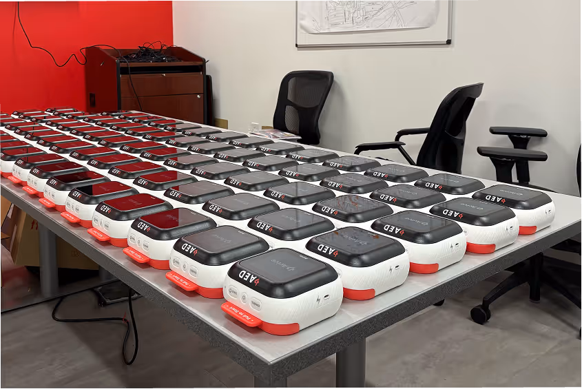 A table filled with multiple Avive AEDs neatly arranged in rows in a room with chairs and a red wall.
