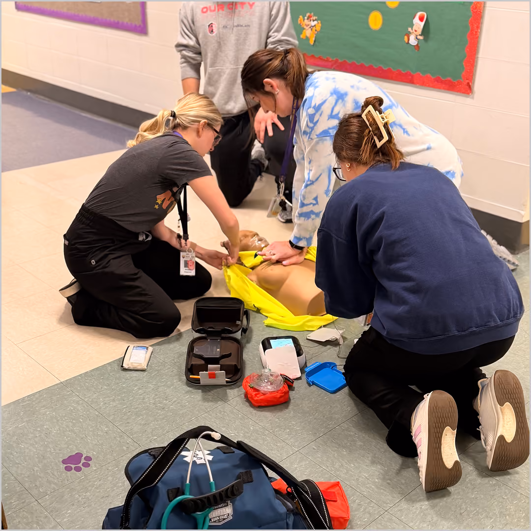 Three people kneeling on a tiled floor performing CPR on a training mannequin wearing a yellow jacket, with medical equipment including a Avive AED and a stethoscope nearby.