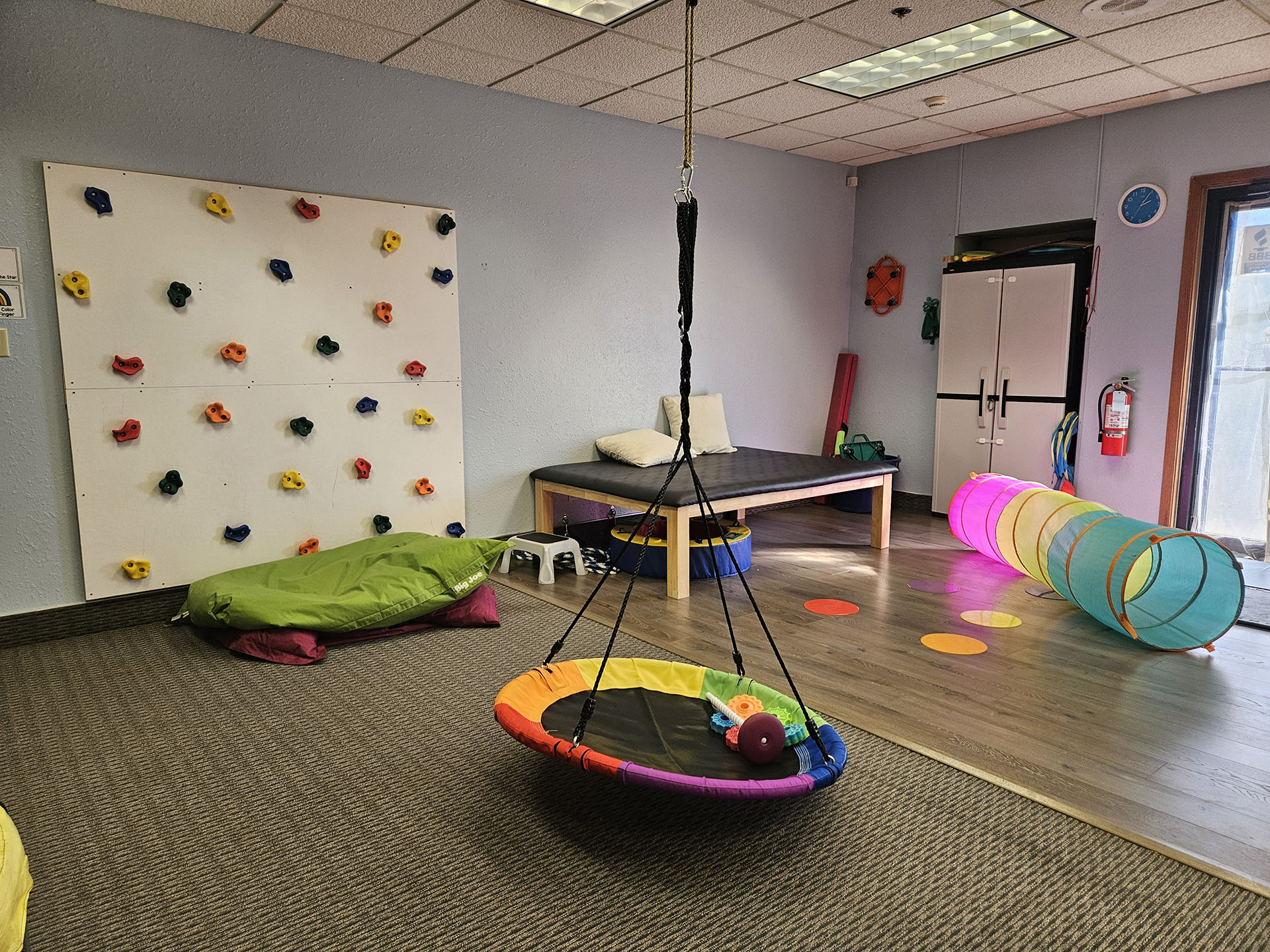 Colorful pediatric therapy room at Advanced Therapy Clinic in Butte, featuring a climbing wall, sensory swing, play tunnel, and cozy seating — designed to support joyful, individualized care for children.