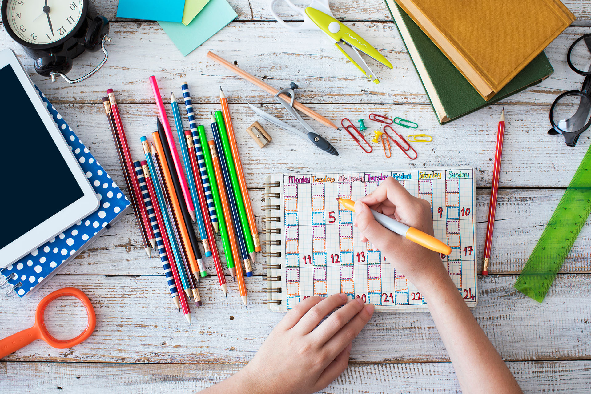 Hand writing in a colorful weekly visual schedule surrounded by school supplies, illustrating how structured routines support children during back‑to‑school transitions.