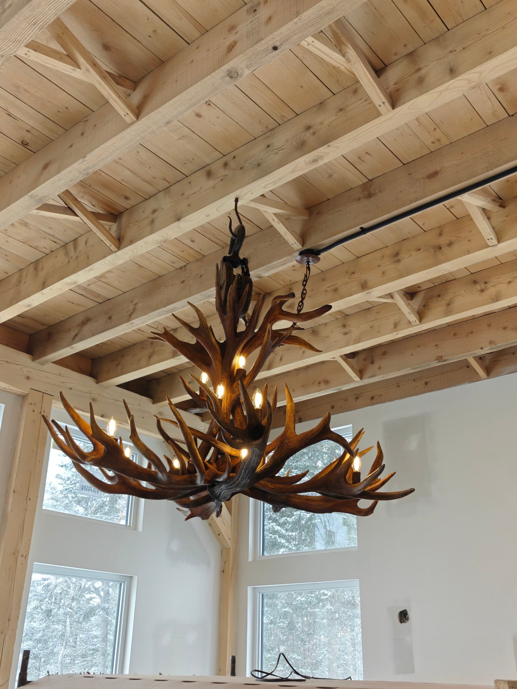 Rustic chandelier made of deer antlers hanging from a wooden ceiling in a room with large windows showing snowy trees outside.