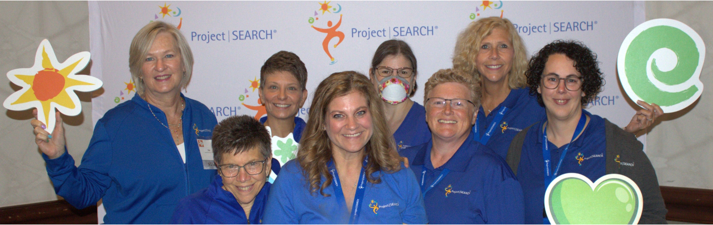 Group of eight individuals wearing matching blue Project SEARCH shirts, standing in front of a Project SEARCH backdrop and holding colorful cut-out shapes like hearts and stars, representing team spirit and program participation.