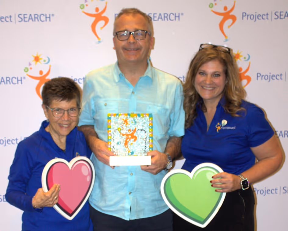 Three individuals stand in front of a backdrop with repeating 'Project SEARCH' logos. The person on the left holds a pink heart sign. The person in the center holds a colorful framed artwork featuring the Project SEARCH logo. The person on the right wears a blue Project SEARCH shirt holding a green heart sign. The image appears to be from an award or recognition event.