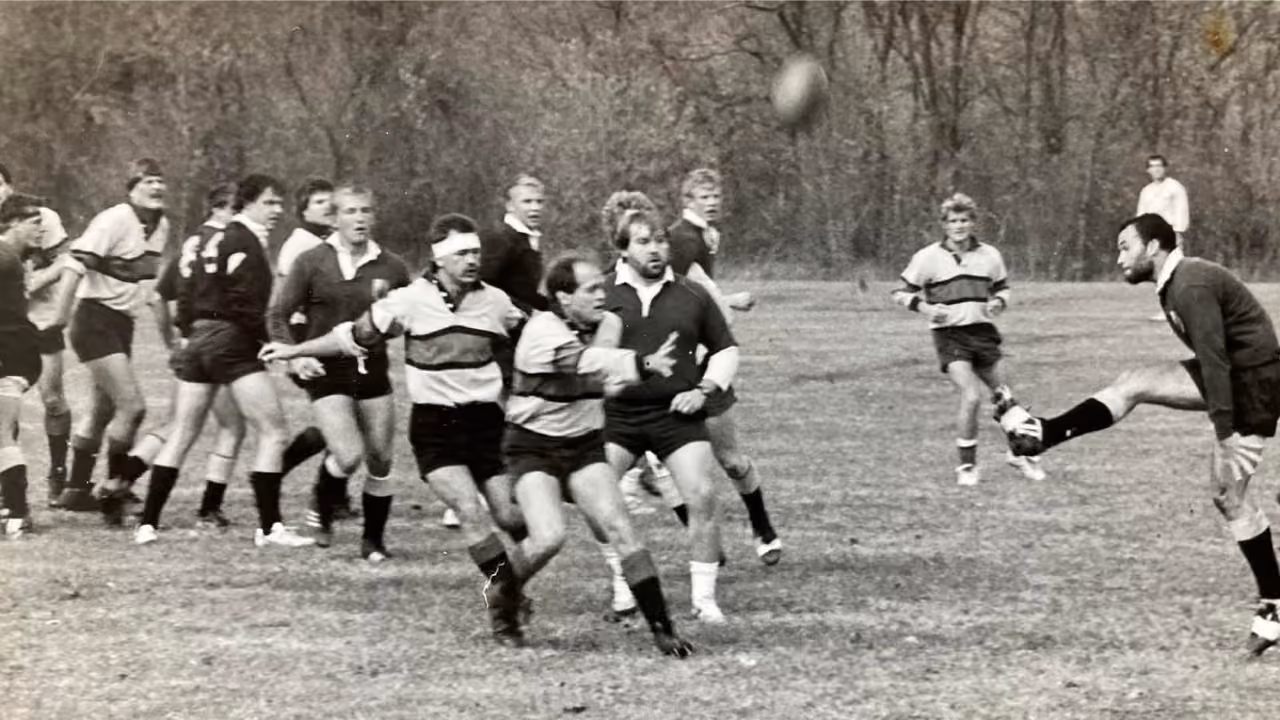 Black and white photo of rugby players in action with ball in mid-air on grassy field.