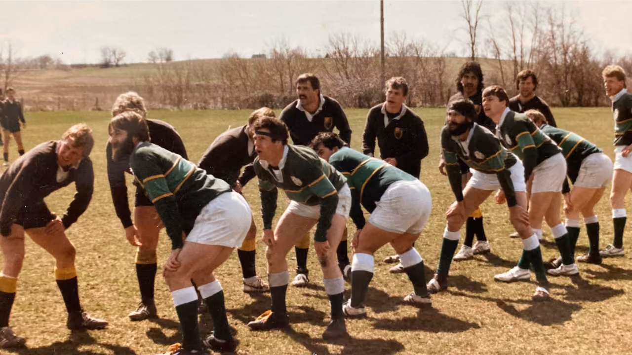 Two rugby teams facing each other in a scrum formation on a grassy field under a cloudy sky.