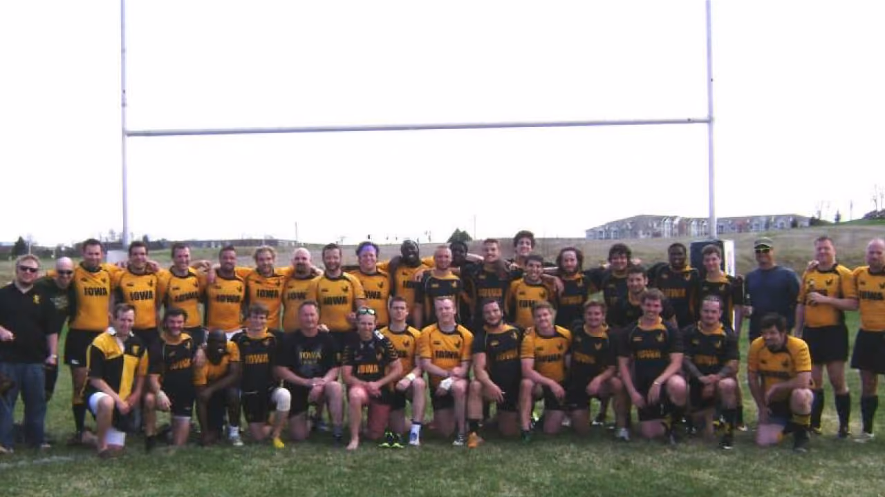 Large group of rugby players in black and yellow Iowa jerseys posing on grass under goalposts.