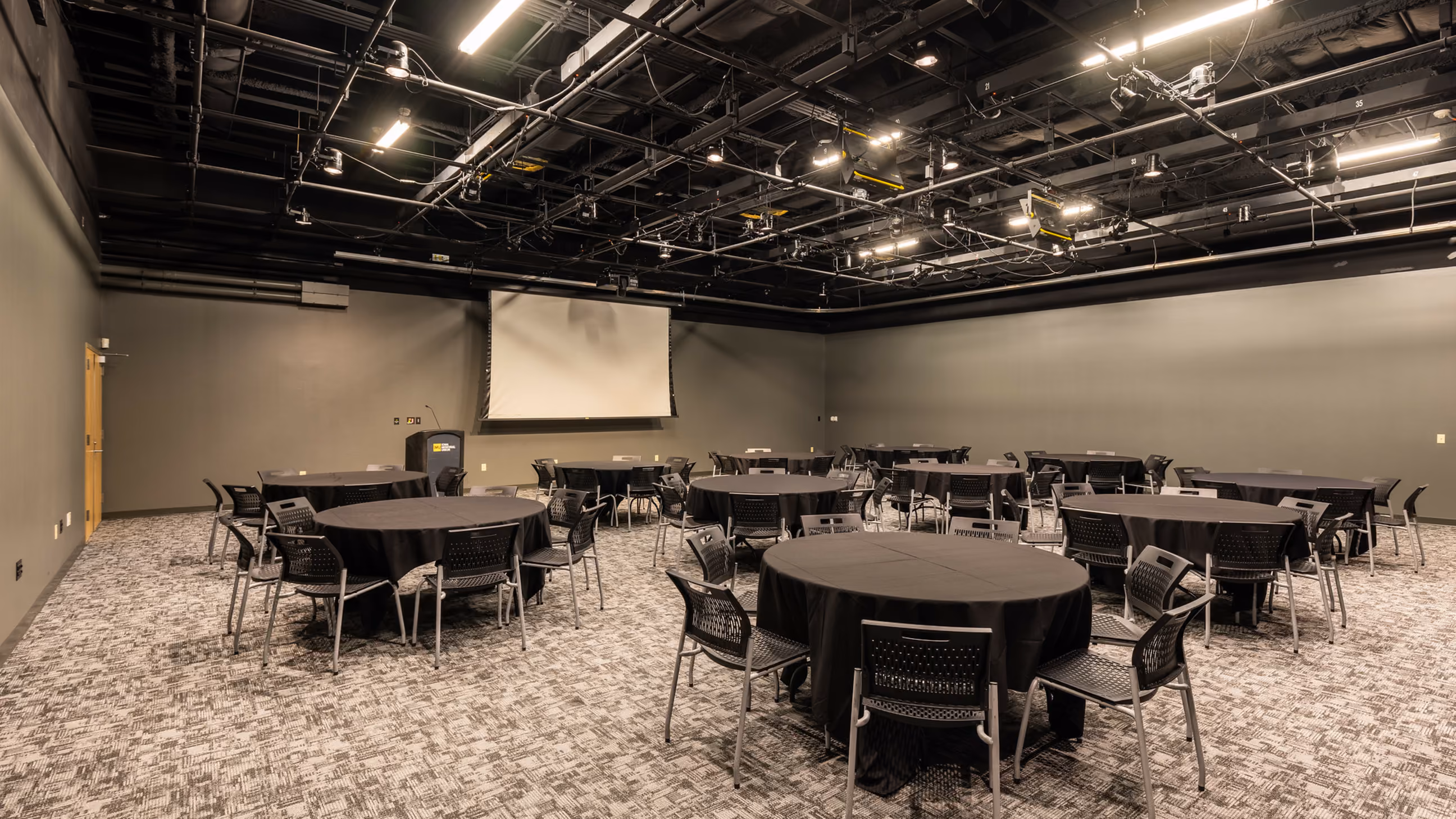 Conference room with round tables covered by black tablecloths and black chairs arranged around them, a podium, a retracted projector screen on the far wall, and ceiling-mounted lighting and cables.