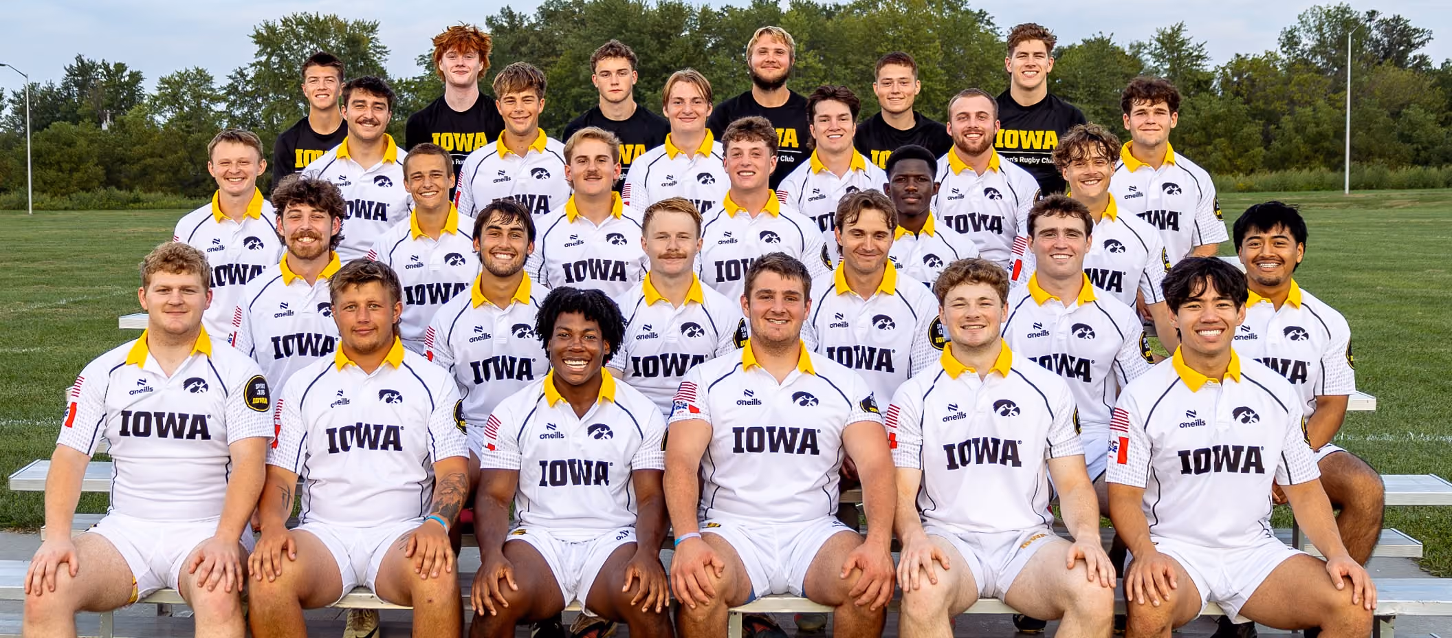 Group photo of 29 male University of Iowa rugby players in white jerseys sitting and standing on bleachers outdoors.