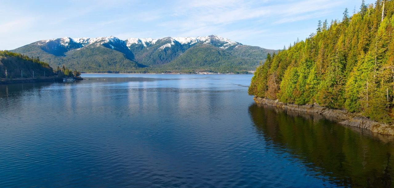 View of calm blue water, lush evergreen coastline, and snow-capped mountains near Ketchikan, Alaska.